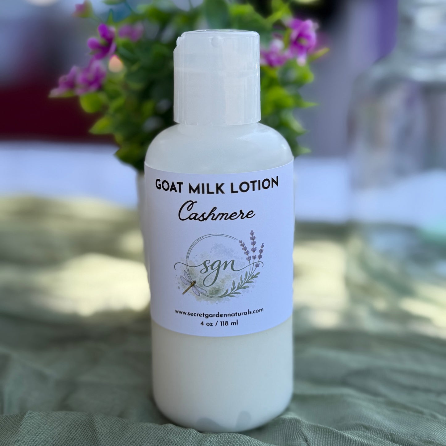 A white bottle of goat milk lotion labeled 'Cashmere' by Secret Garden Naturals, placed on a textured green cloth with blurred purple flowers in the background.