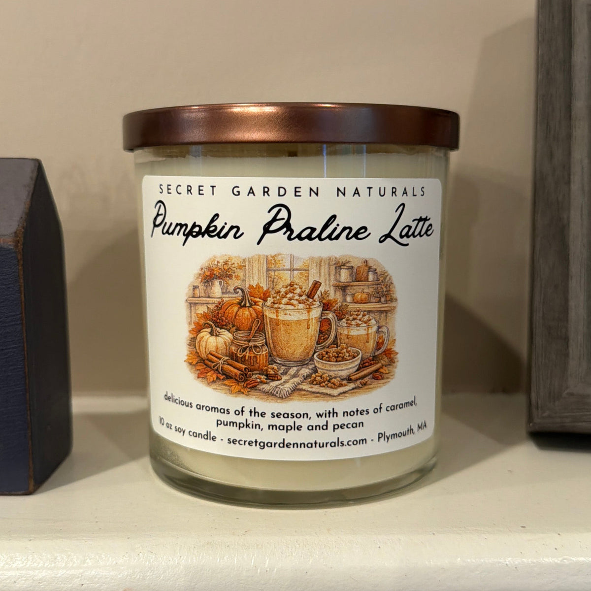 A jar candle labeled 'Pumpkin Praline Latte' by Secret Garden Naturals sits on a white mantel, flanked by a dark blue decorative house and a framed picture on a beige wall.