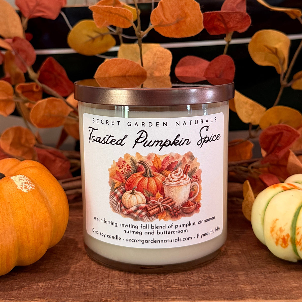 A jar candle labeled 'Toasted Pumpkin Spice' by Secret Garden Naturals sits on a wooden surface, styled with small pumpkins and autumn leaves.