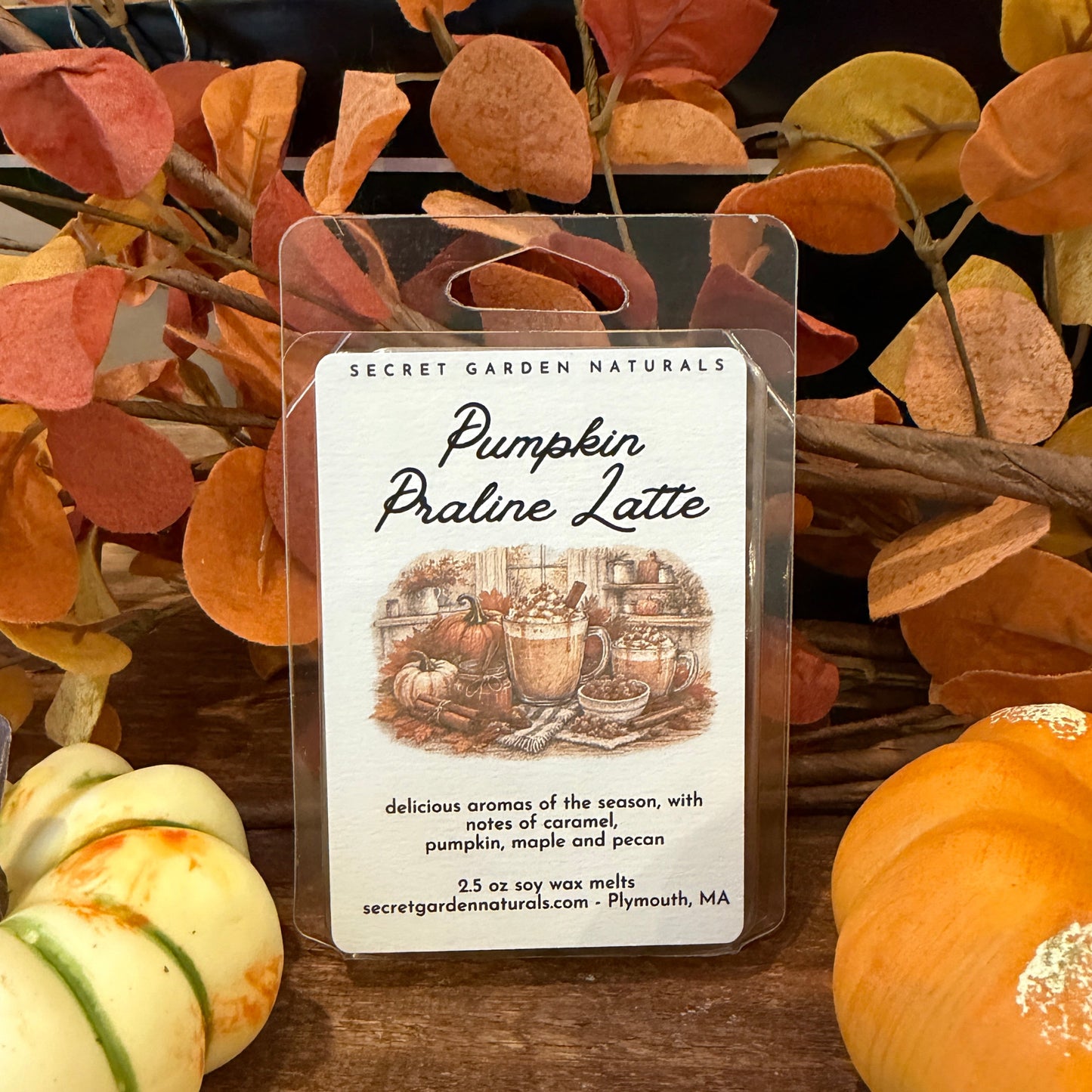A wax melt package labeled 'Pumpkin Praline Latte' by Secret Garden Naturals, displayed in a clear plastic container on a wooden surface with autumn leaves and small pumpkins around it.