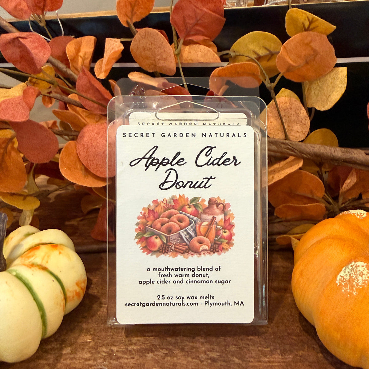 A clear plastic clamshell package labeled 'Secret Garden Naturals Apple Cider Donut' sits on a wooden surface, surrounded by autumn leaves, pumpkins, and branches.