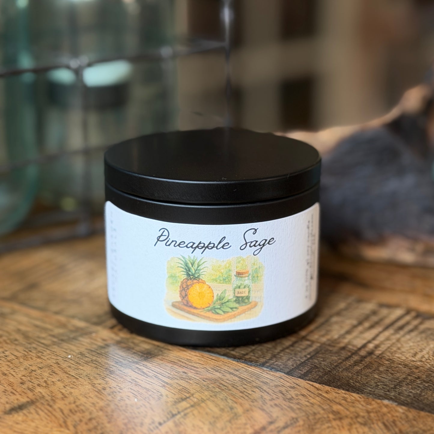 Black candle tin labeled 'Pineapple Sage' on a wooden surface with a blurred background