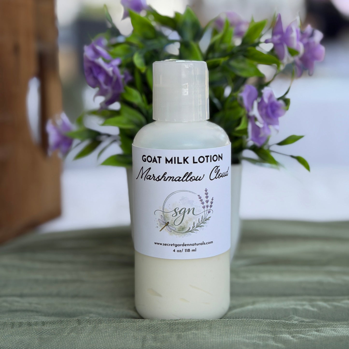 A bottle of Secret Garden Naturals Goat Milk Lotion in Marshmallow Cloud, filled with white lotion, set on a green fabric surface with a blurred background of purple flowers and a wooden structure.