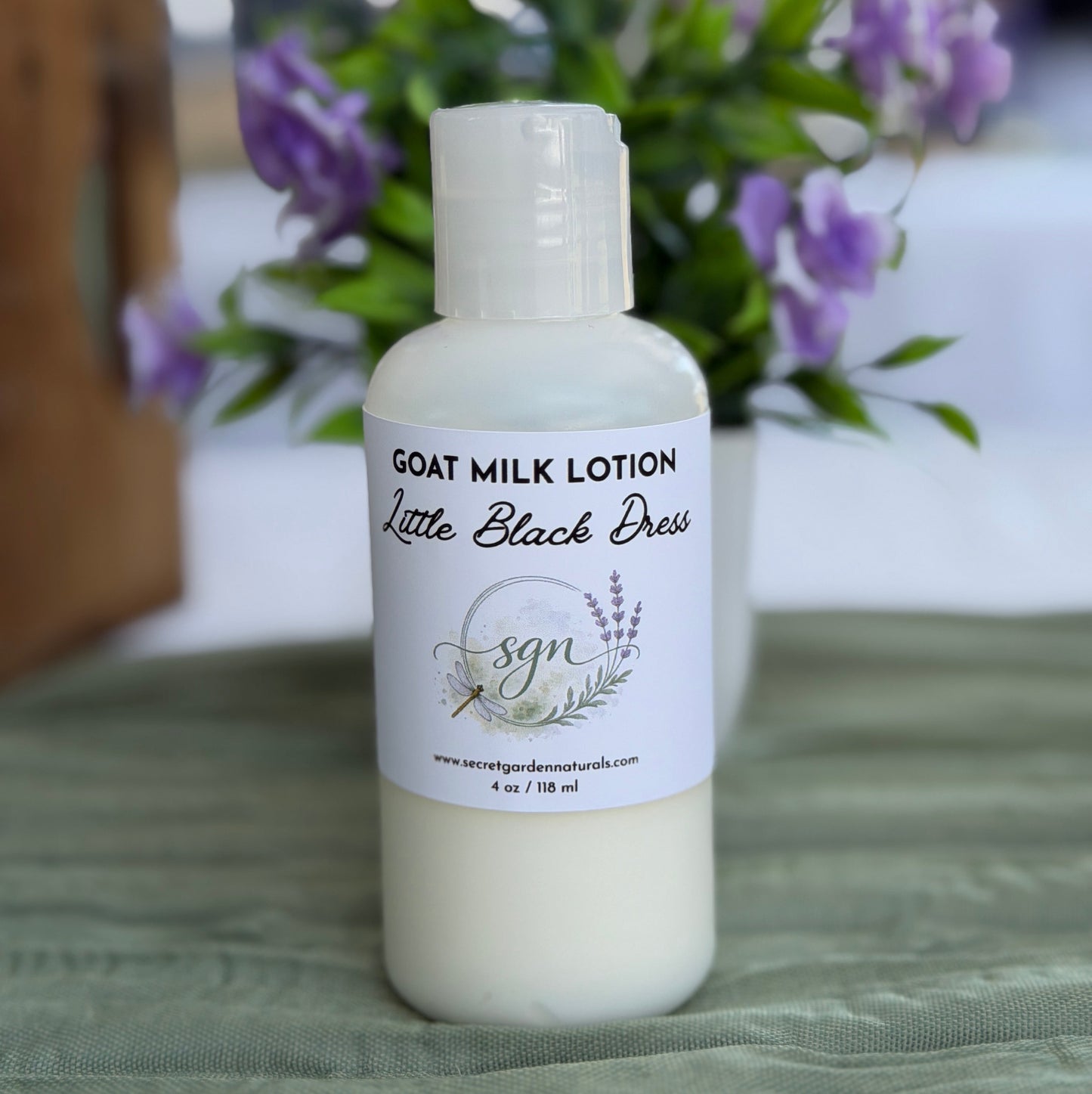 A bottle of goat milk lotion by Secret Garden Naturals titled Little Black Dress sits on a textured green cloth with a blurred background of purple flowers.