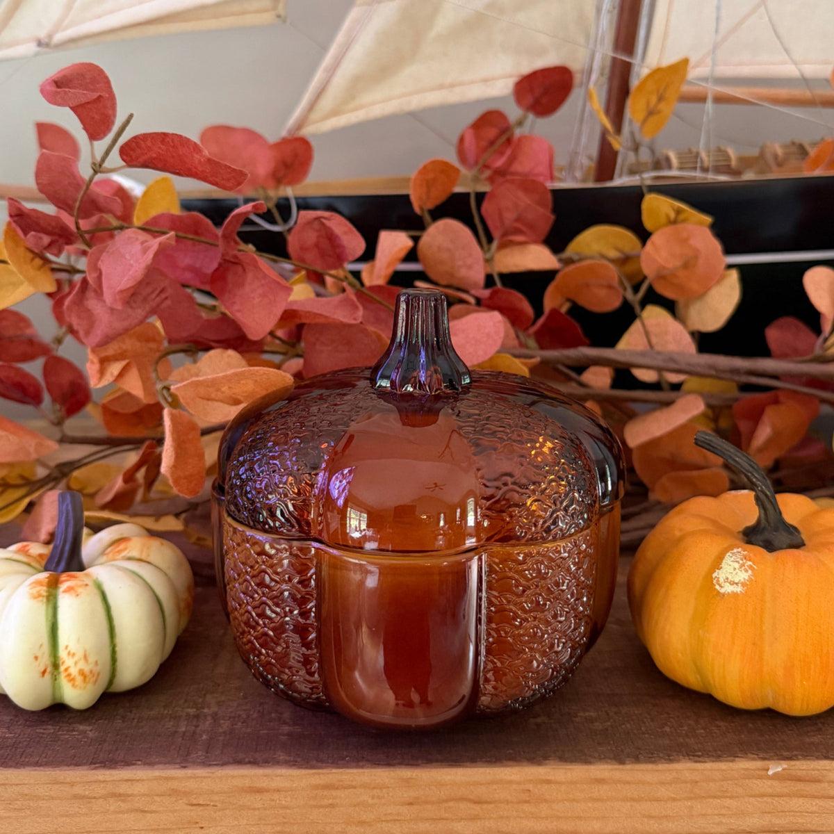 An amber glass pumpkin-shaped container with a textured pattern, styled on a wooden surface with autumn leaves, a small white pumpkin, and an orange pumpkin, with a model sailboat in the background.