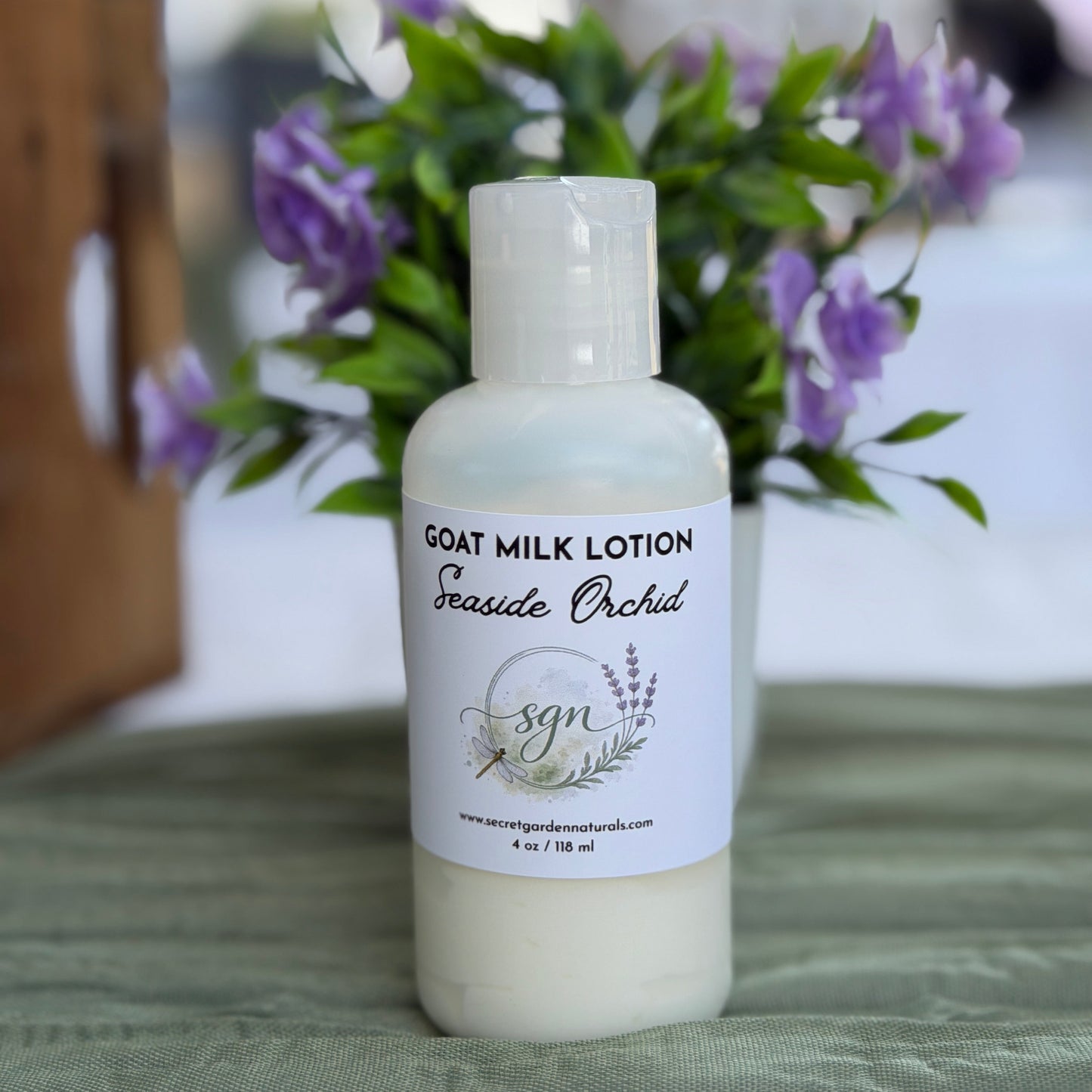 A bottle of Seaside Orchid Goat Milk Lotion by Secret Garden Naturals sits on a green textured cloth with a blurred background of purple flowers and a wooden surface.