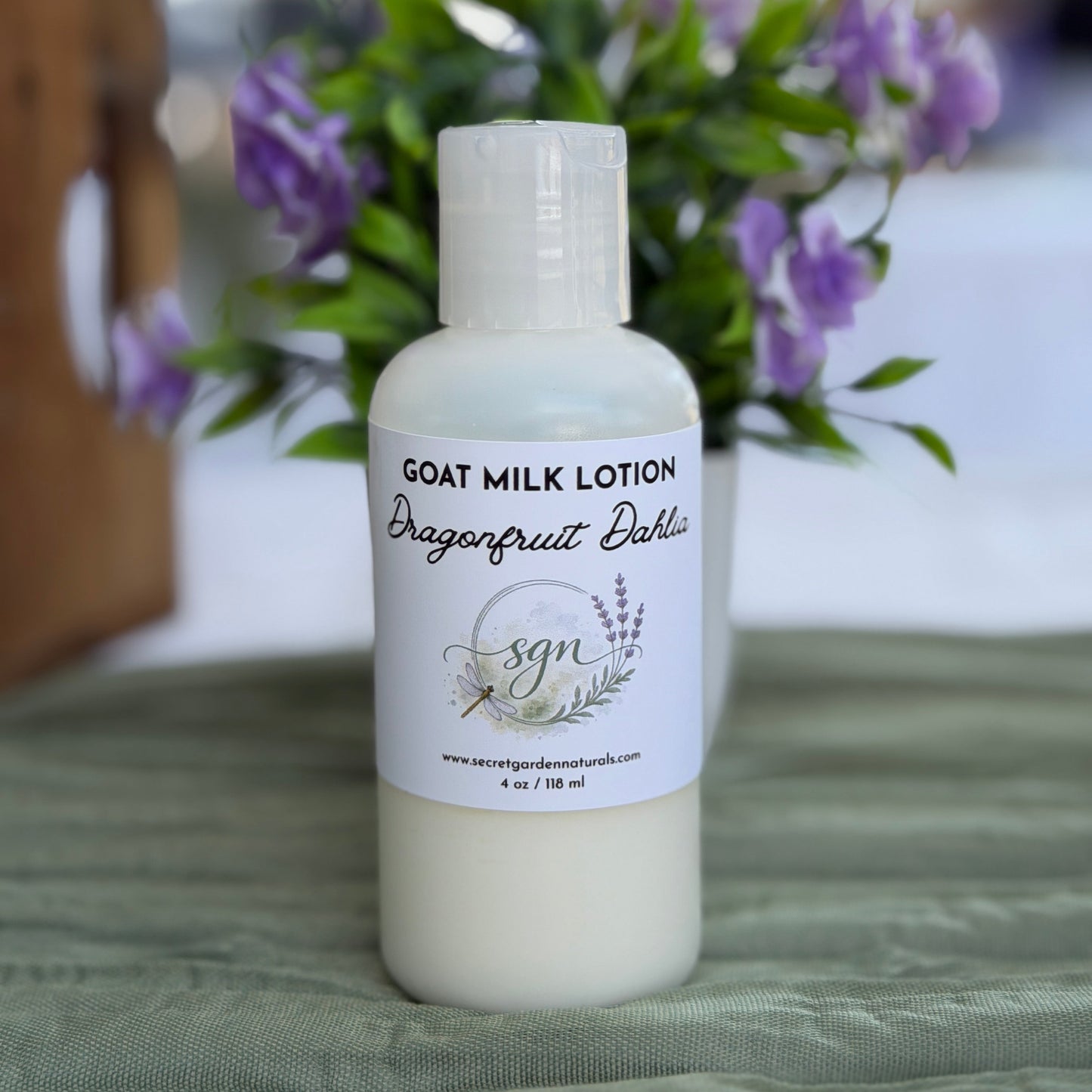 A 4 oz bottle of goat milk lotion with a white cap and white label, 'Dragonfruit Dahlia' scent and initials 'sgn', set on a green fabric surface with blurred purple flowers in the background.