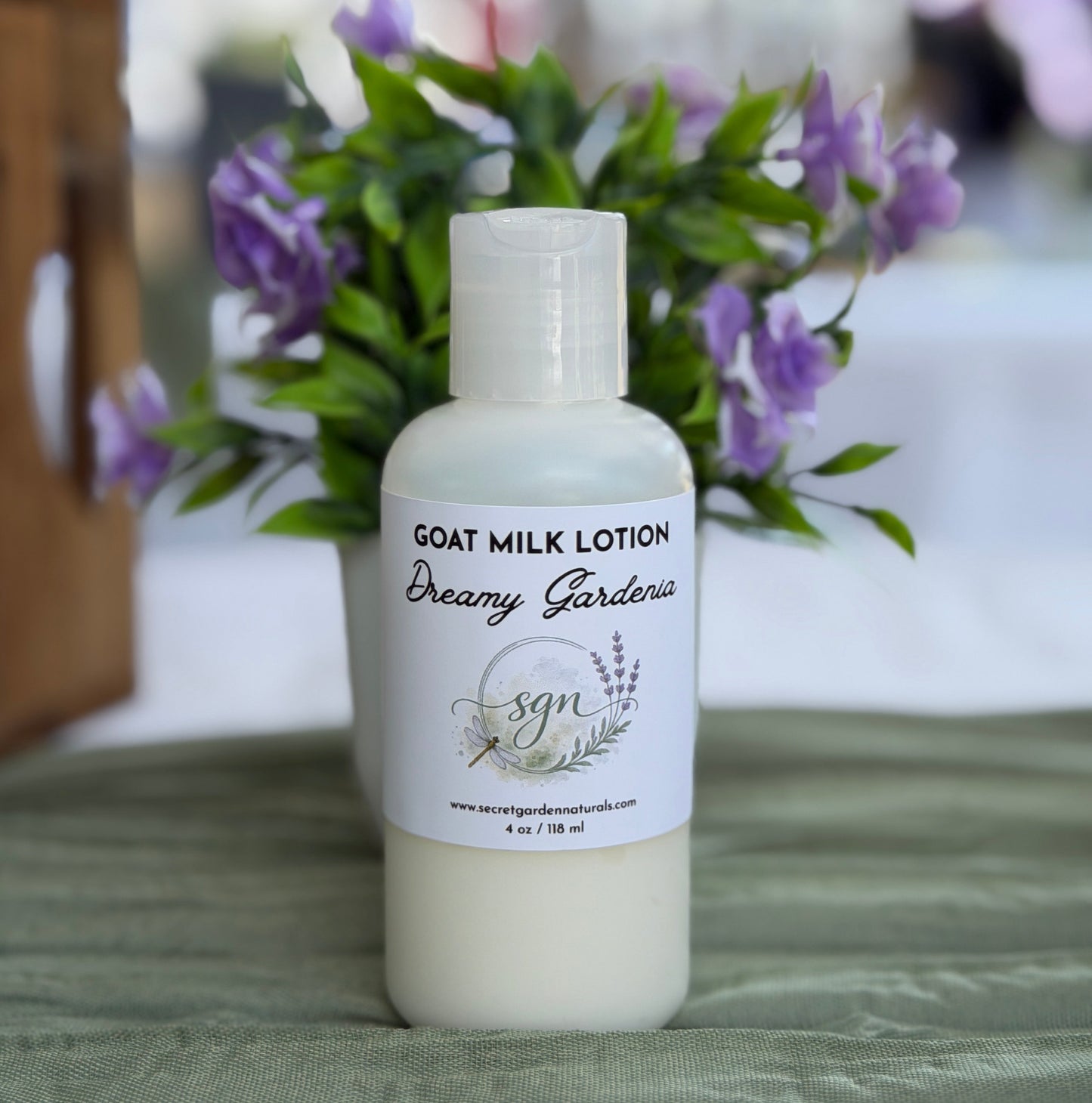 A bottle of Dreamy Gardenia Goat Milk Lotion by sgn, set on a green textured cloth with a blurred background of purple flowers and greenery.