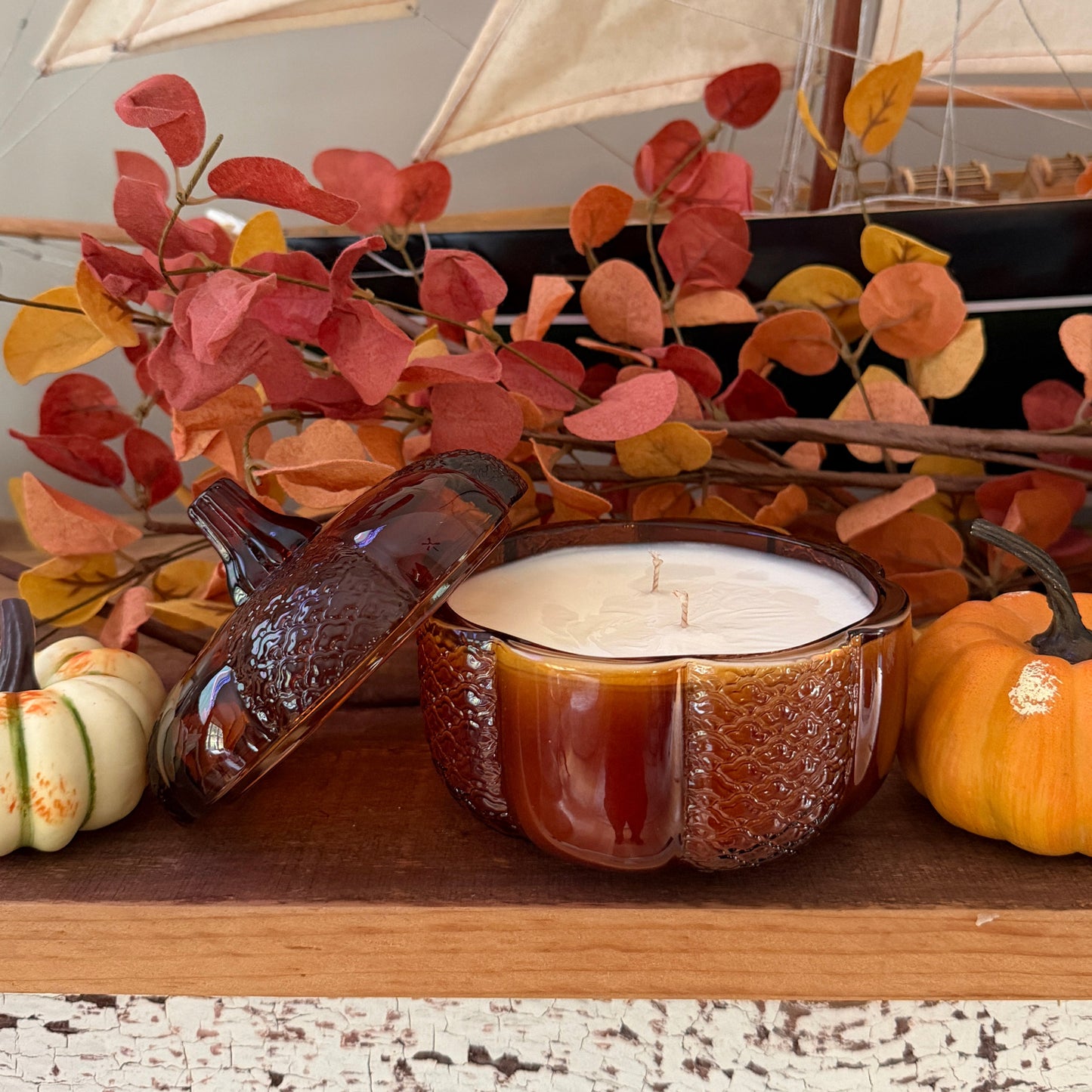 A  candle in a amber glass pumpkin jar with its lid leaning beside it, styled on a wooden surface with miniature pumpkins and  autumn leaves.
