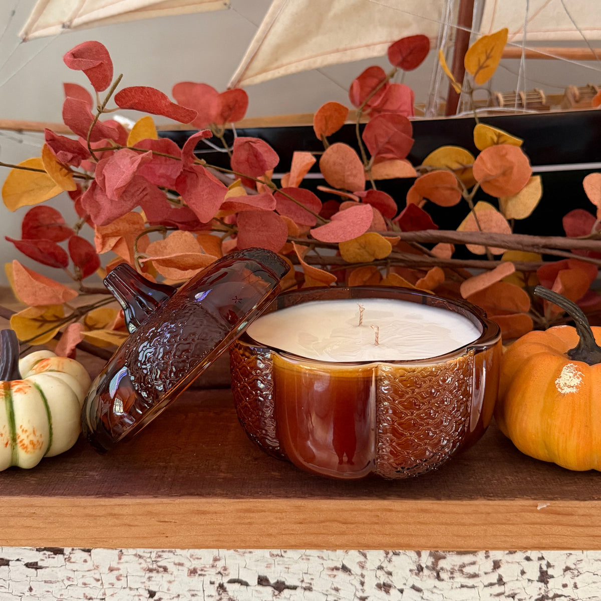 A  candle in a amber glass pumpkin jar with its lid leaning beside it, styled on a wooden surface with miniature pumpkins and  autumn leaves.