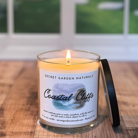 Candle labeled 'Coastal Cliffs' from Secret Garden Naturals on a wooden surface with a blurred outdoor background.