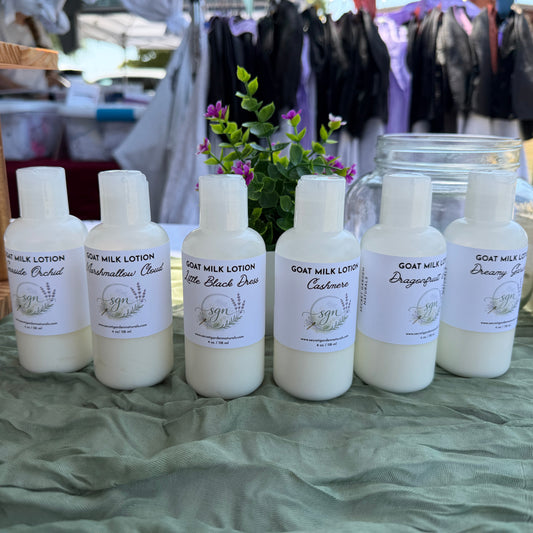 Six small white bottles of goat milk lotion with white labels are lined up on a green cloth-covered table at an outdoor market. The labels show different scents including Seaside Orchid, Marshmallow Cloud, Little Black Dress, Cashmere, Dragonfruit Dahlia, and Dreamy Gardenia.