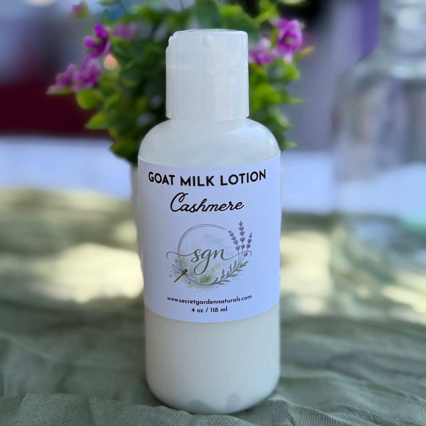 A bottle of Secret Garden Naturals Goat Milk Lotion in the 'Cashmere' scent, shown on a textured green cloth with a blurred background of purple flowers.