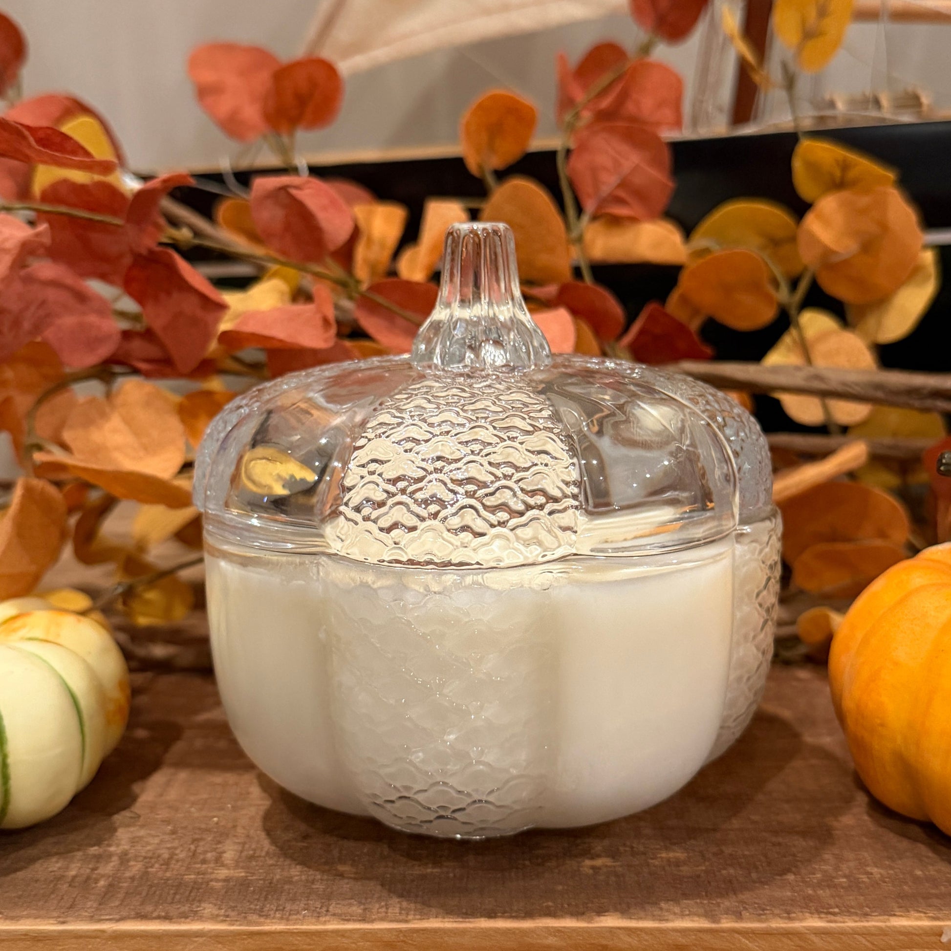 A candle in a clear glass pumpkin shaped jar on a wooden shelf with autumn leaves and pumpkins.