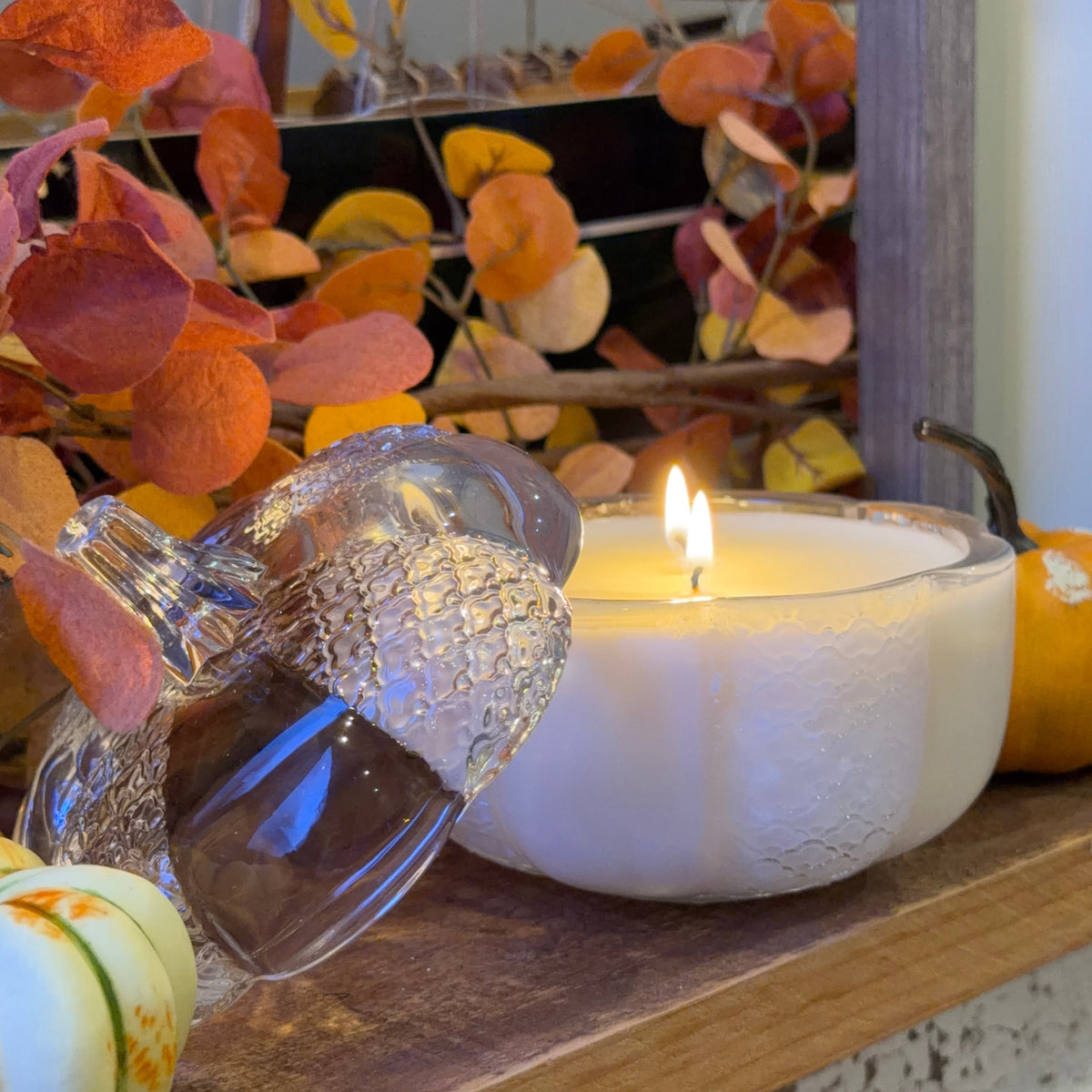 A photo of a double wicked lit candle in a clear glass pumpkin jar on a wooden shelf with pumpkins and autumn leaves.