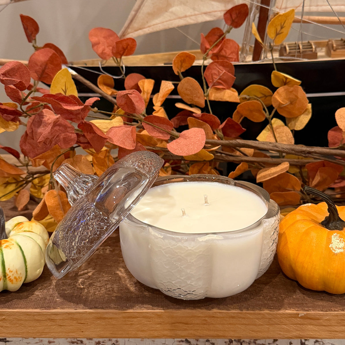 A double wicked candle in a clear glass pumpkin shaped jar on a shelf with pumpkins and autumn leaves.