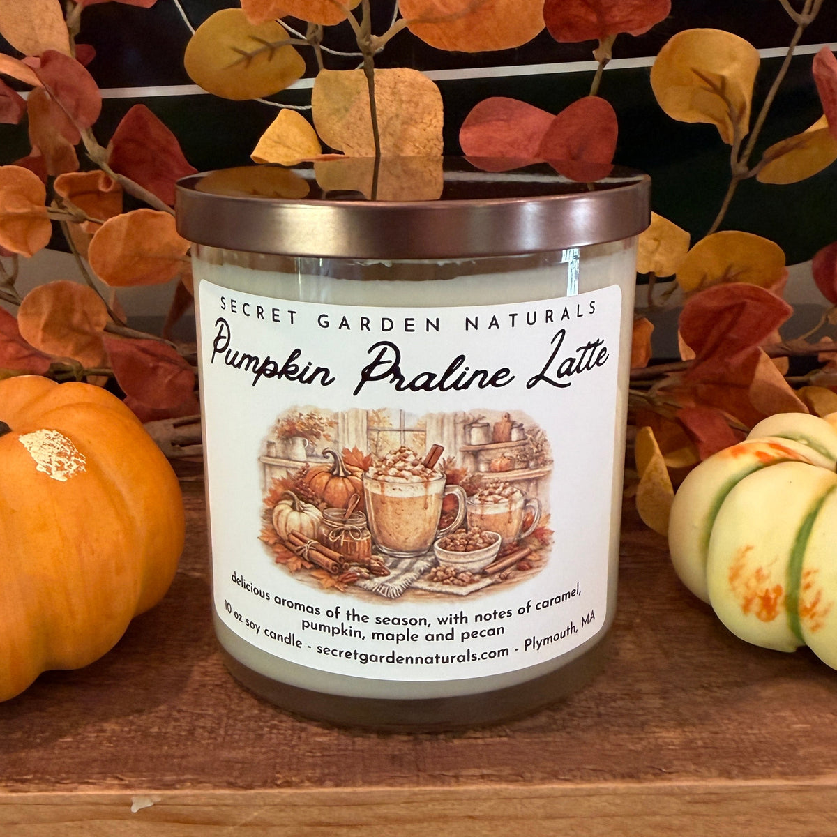 A jar candle labeled 'Pumpkin Praline Latte' by Secret Garden Naturals, styled on a wooden shelf with decorative pumpkins and autumn leaves.