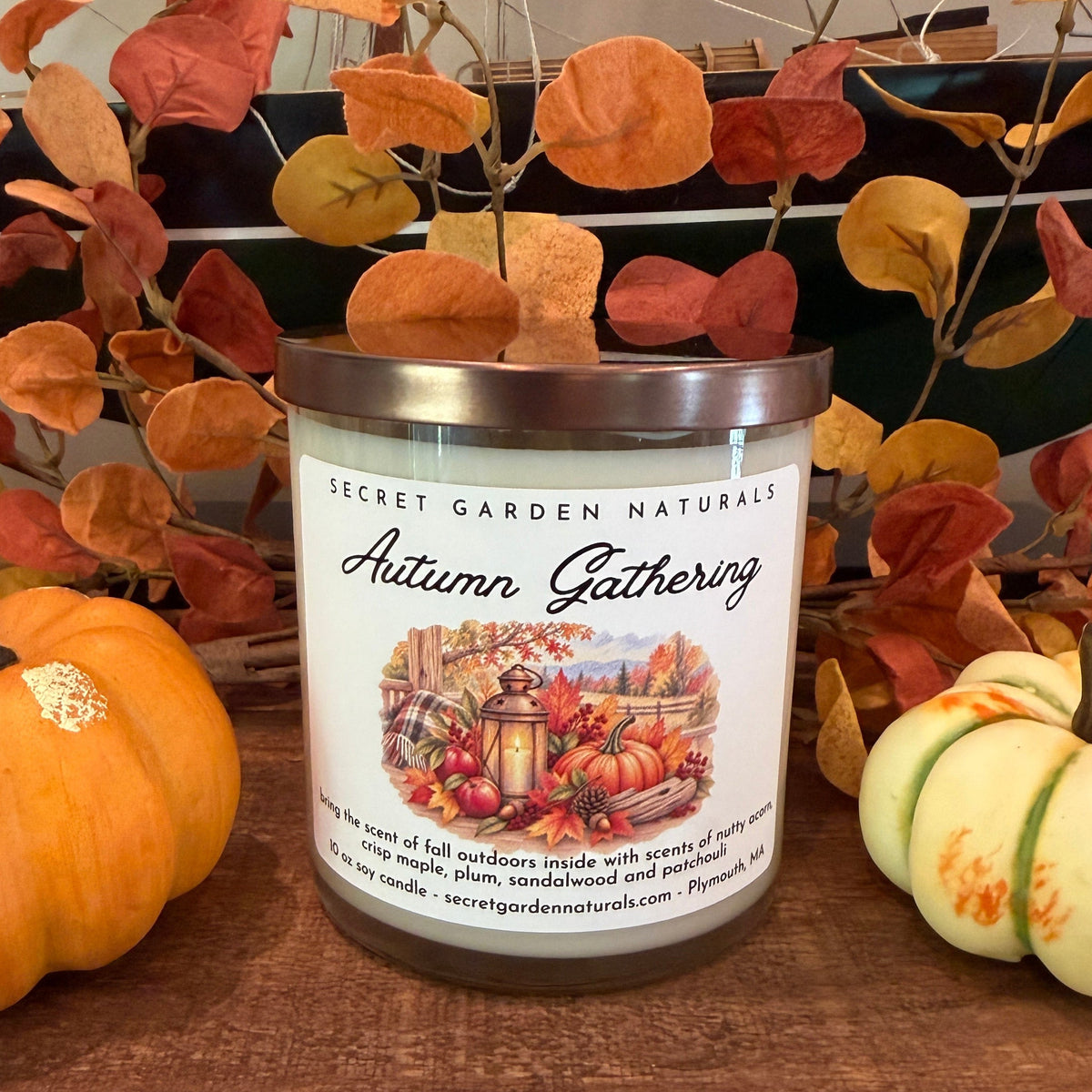 A jar candle labeled 'Secret Garden Naturals Autumn Gathering' sits on a wooden surface, styled with small pumpkins and fall leaves in the background.