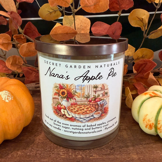 A jar candle labeled 'Nana's Apple Pie' by Secret Garden Naturals sits on a wooden surface, styled with small pumpkins and autumn leaves in front of a ship model.