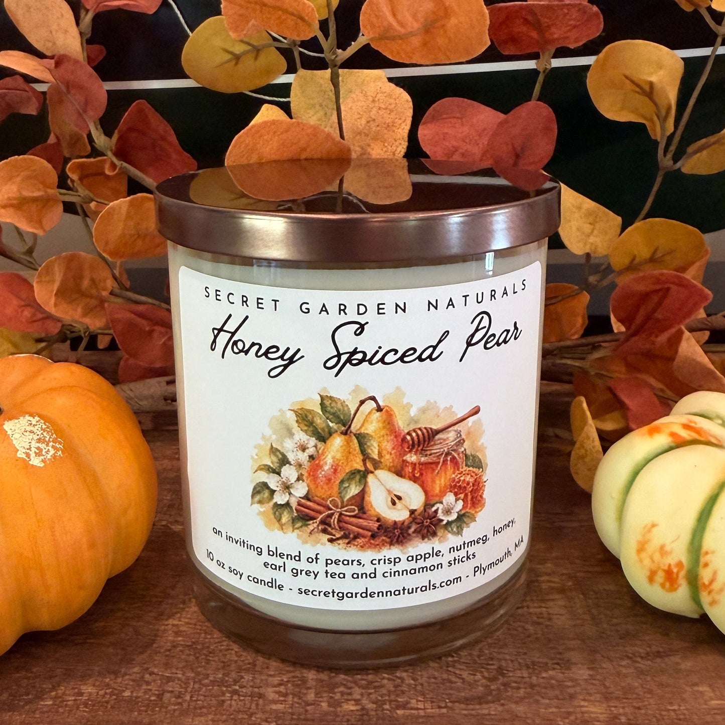 A jar candle labeled 'Secret Garden Naturals Honey Spiced Pear' sits on a wooden surface, styled with small pumpkins and autumn leaves in front of a ship model.