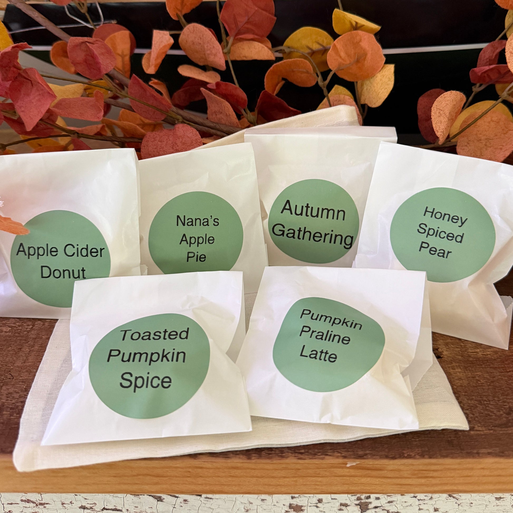 Six white paper gift bags with green circular labels displaying autumn-themed scents and names, arranged on a wooden surface with fall leaves in the background.