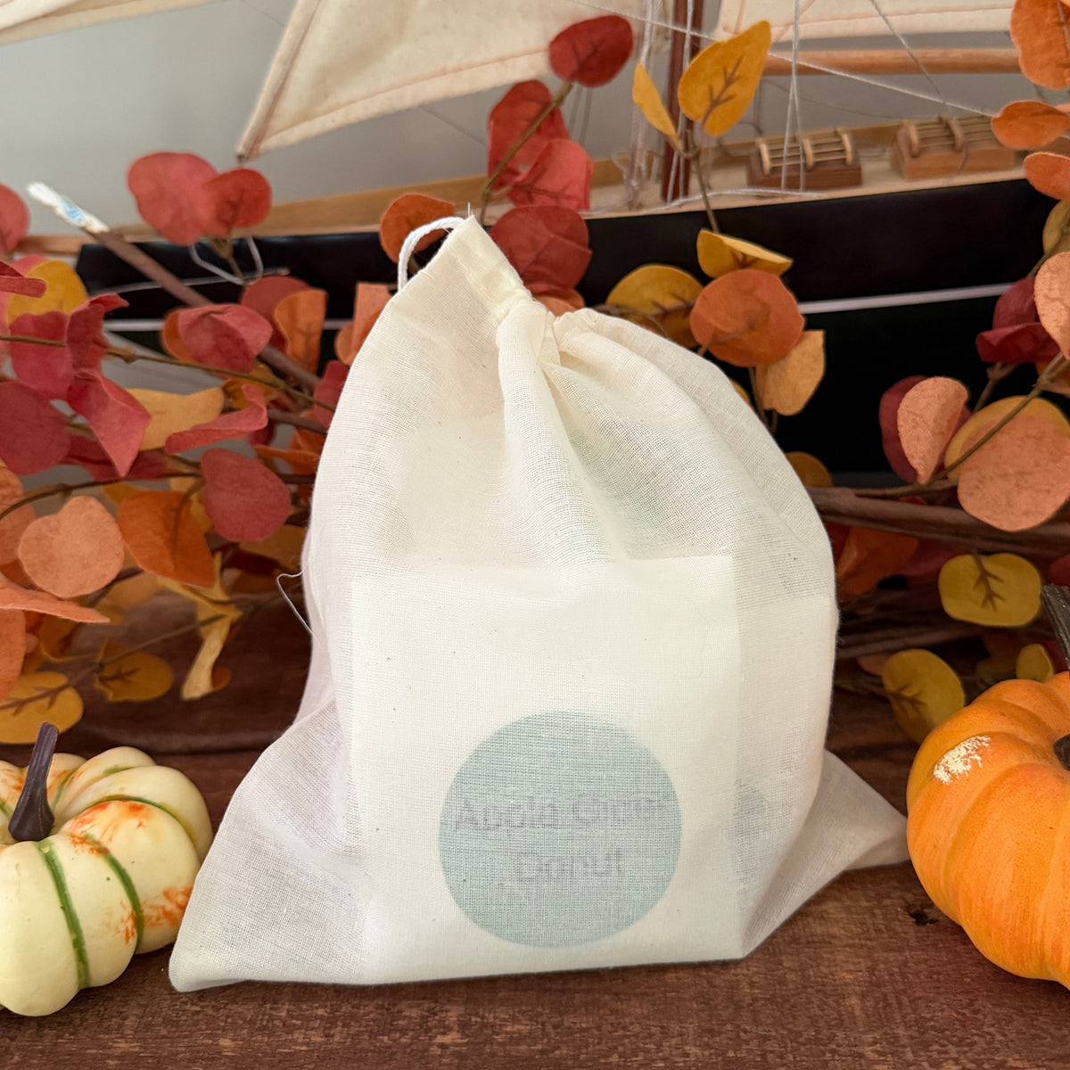 A white drawstring bag containing samples of each of the 6 fall seasonal scents, styled on a wooden surface with autumn leaves and small pumpkins in the background.