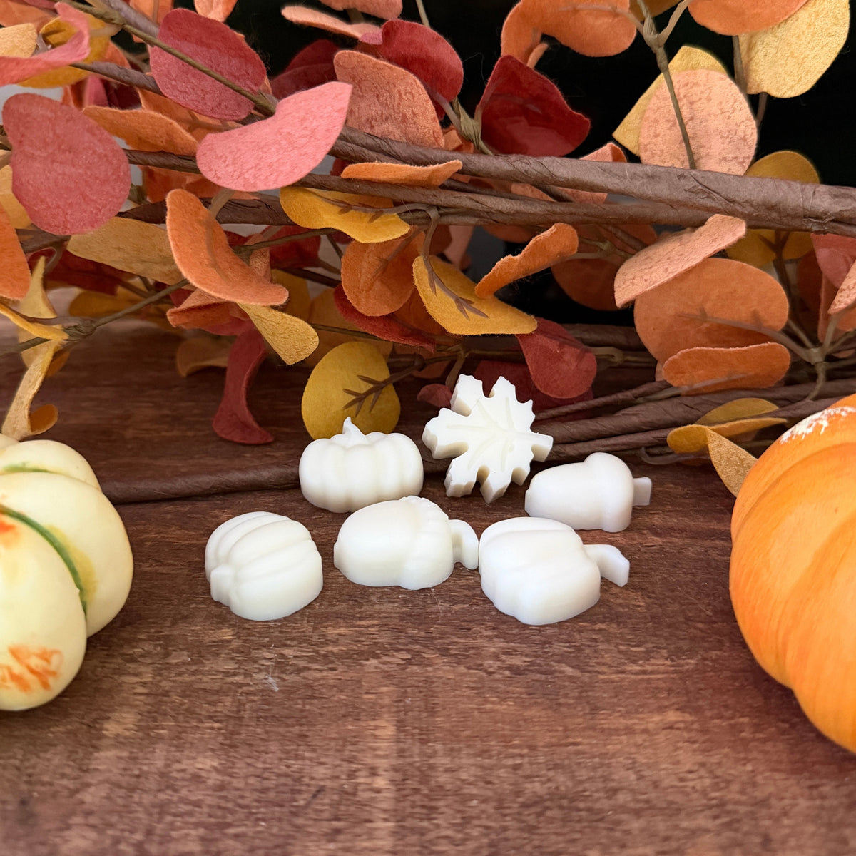 A set of white wax melts in autumn-themed shapes, including acorn, pumpkin, and snowflake designs, arranged on a wooden surface with decorative fall leaves and small pumpkins.