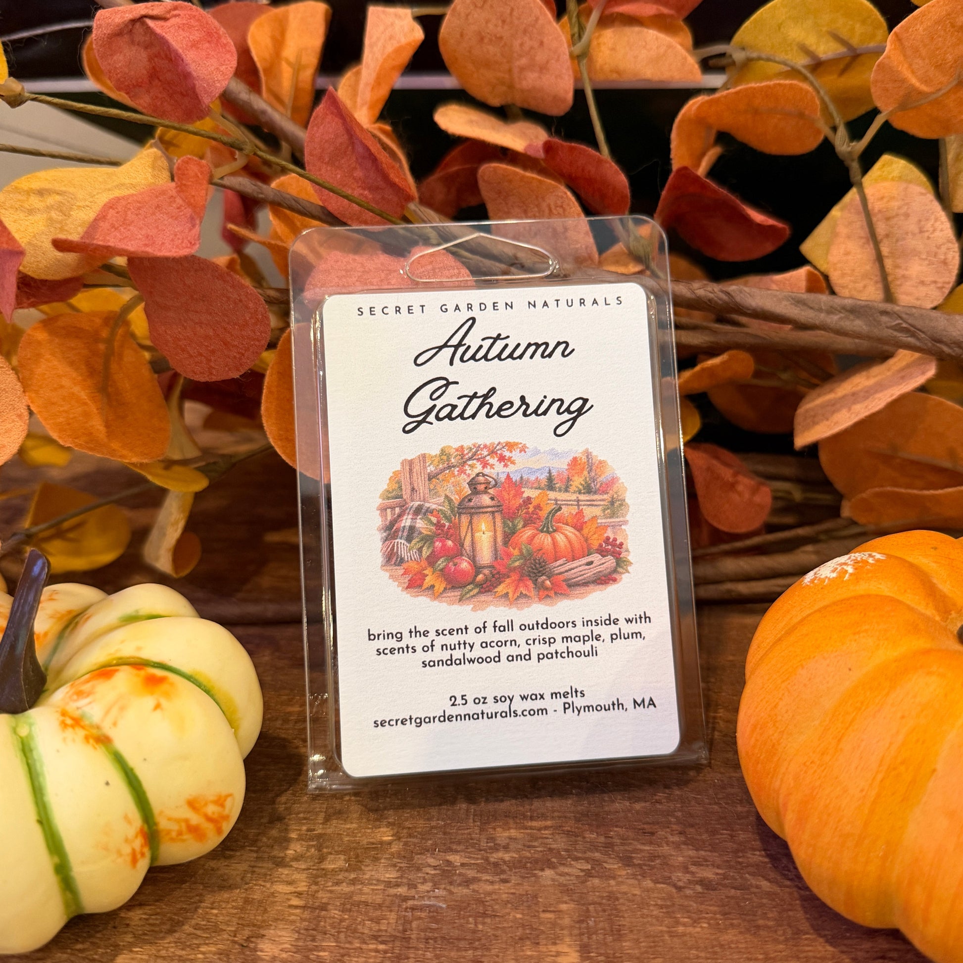 An 'Autumn Gathering' scented wax melt in a clear plastic clamshell package, displayed on a wooden table with decorative autumn leaves, small pumpkins, and a black tray in the background.