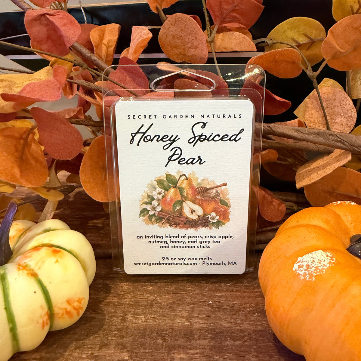 A clear plastic clamshell wax melt package labeled 'Secret Garden Naturals Honey Spiced Pear' stands on a wooden surface surrounded by autumn leaves and small pumpkins.