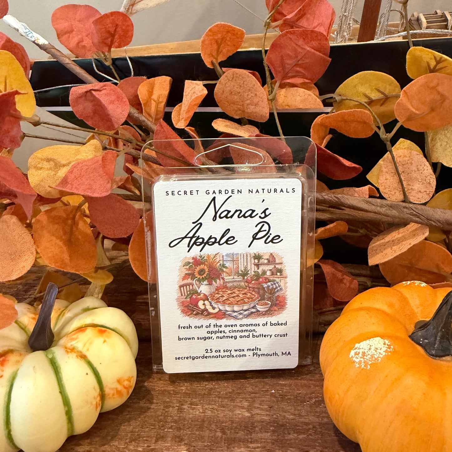 A packaged wax melt labeled 'Nana's Apple Pie' by Secret Garden Naturals, displayed on a wooden surface with autumn leaves, small pumpkins, and a sailboat decoration in the background.