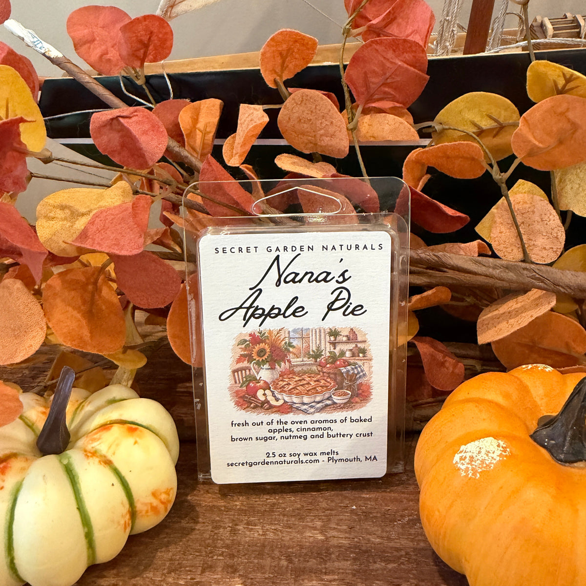 A packaged wax melt labeled 'Nana's Apple Pie' by Secret Garden Naturals, displayed on a wooden surface with autumn leaves, small pumpkins, and a sailboat decoration in the background.