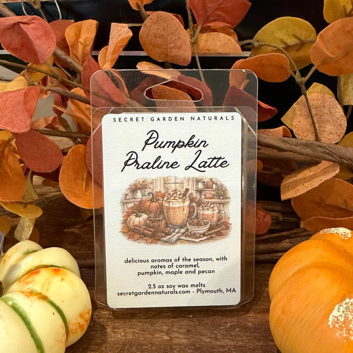 A wax melt package labeled 'Pumpkin Praline Latte' by Secret Garden Naturals, displayed in a clear plastic container on a wooden surface with autumn leaves and small pumpkins around it.