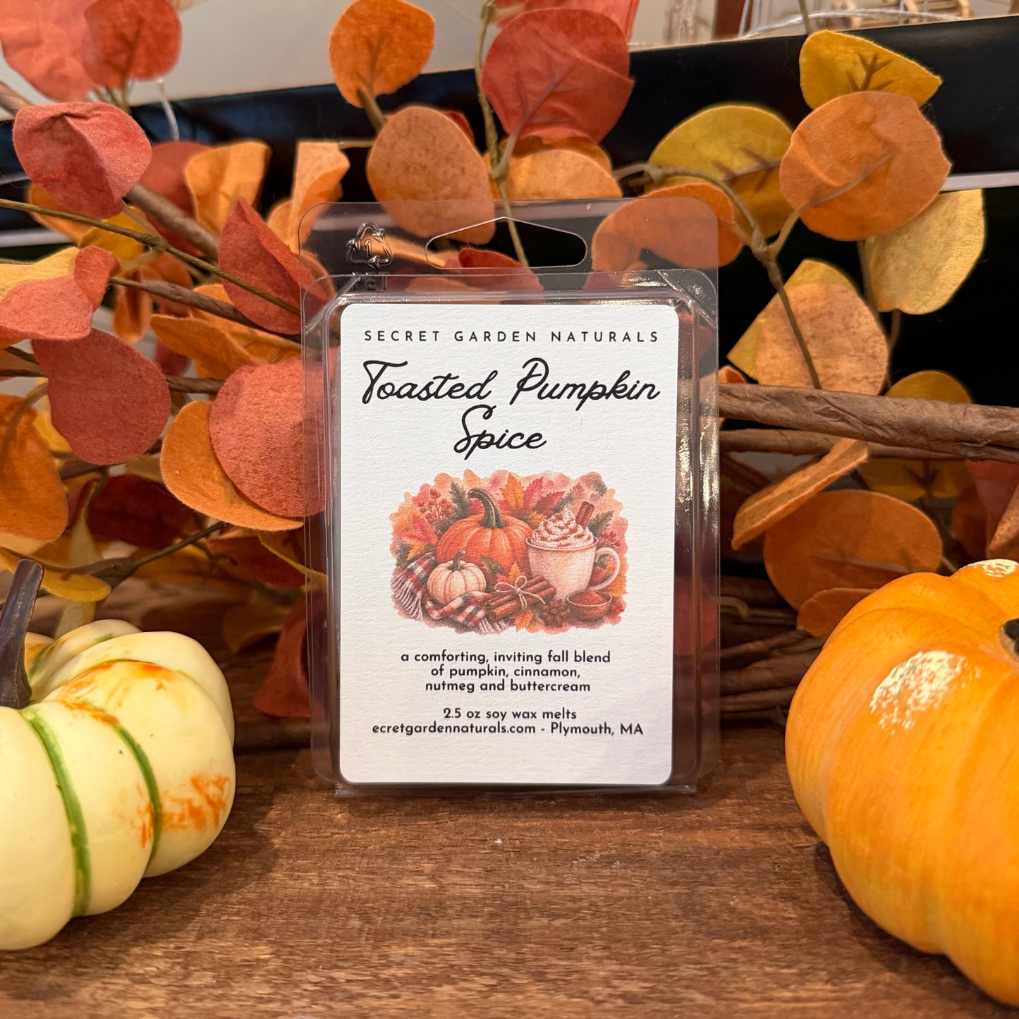 A wax melt package labeled 'Secret Garden Naturals Toasted Pumpkin Spice' stands on a wooden surface, surrounded by fall decorations including orange and yellow leaves and small pumpkins..