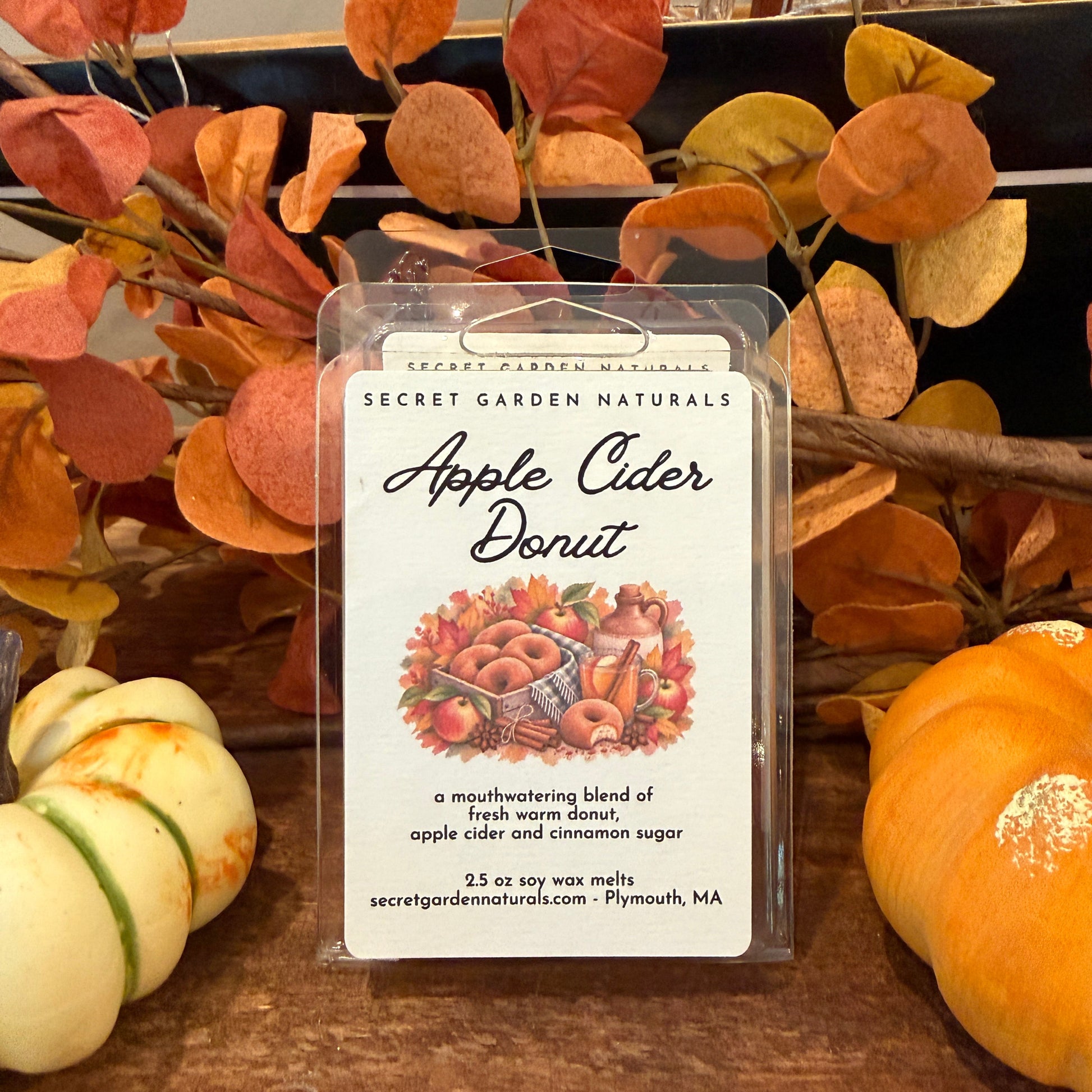 A clear plastic clamshell package labeled 'Secret Garden Naturals Apple Cider Donut' sits on a wooden surface, surrounded by autumn leaves, pumpkins, and branches.