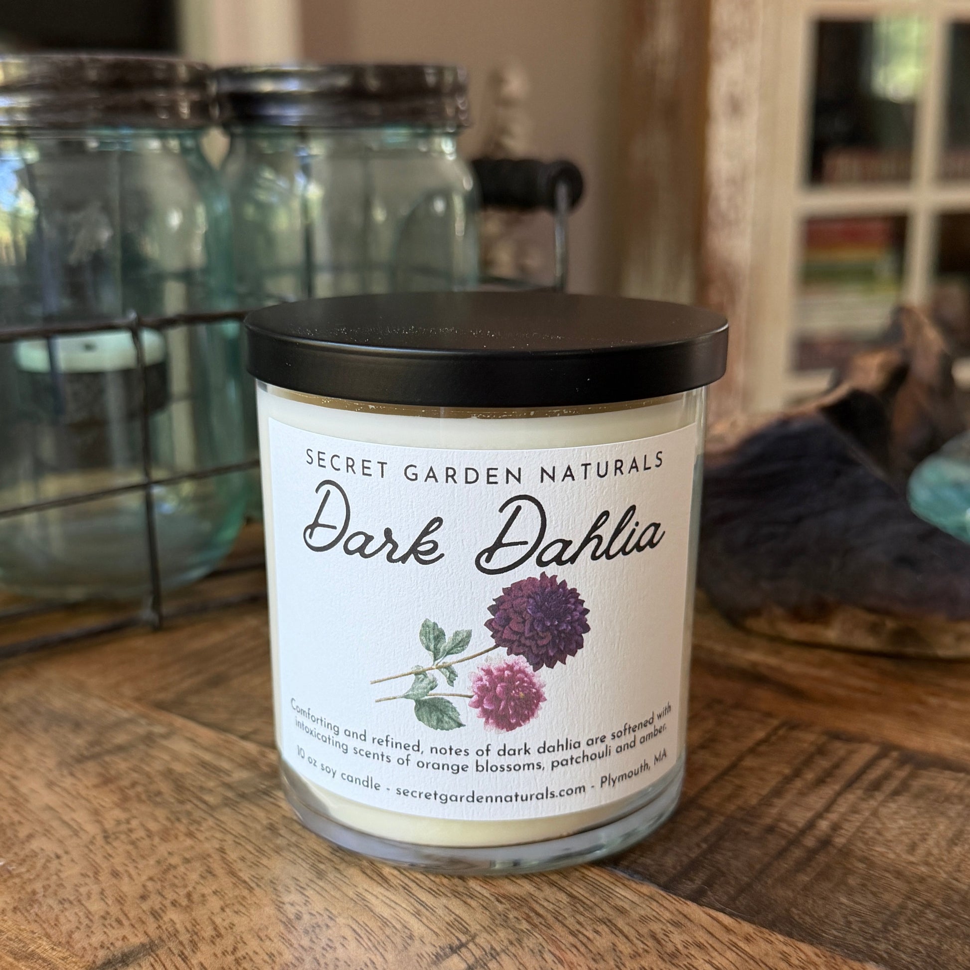 Dark Dahlia candle on a wood table with jars and a cabinet behind it.