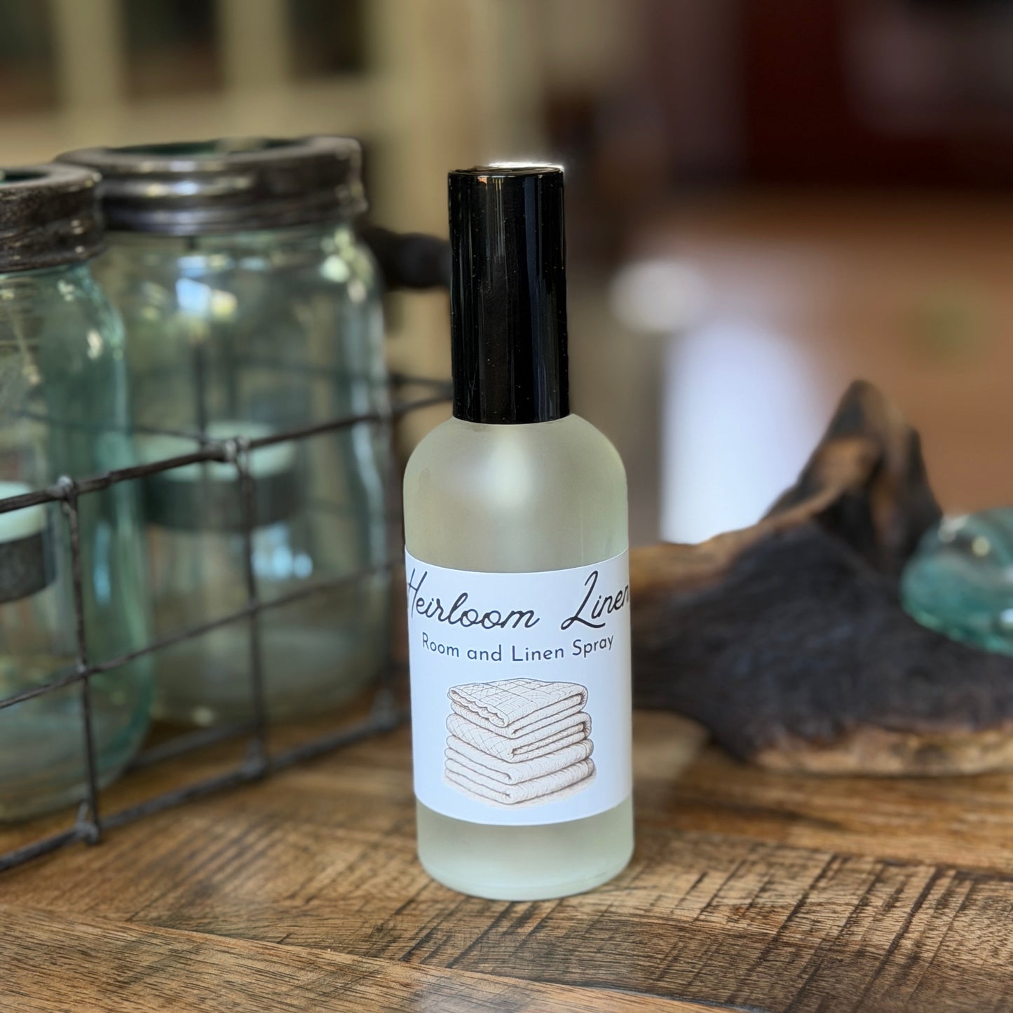Bottle of 'Heirloom Linen' room and linen spray  on a wooden surface with jars in the background
