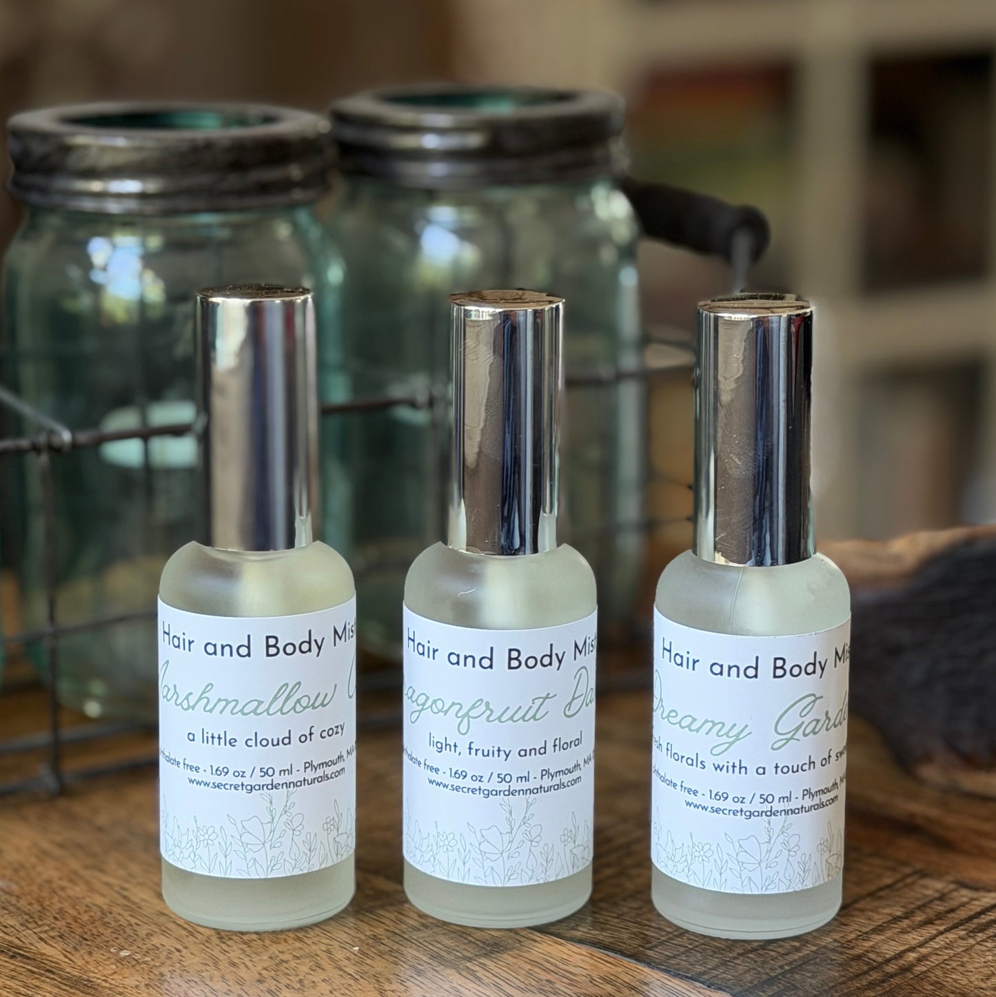 3 glass spray bottles of Secret Garden Naturals Hair and Body Mist on a wooden surface with jars behind them.