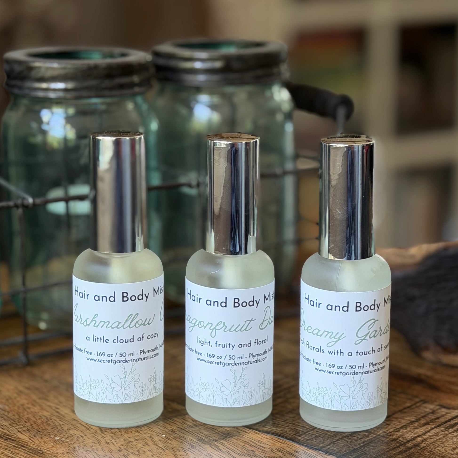 3 glass spray bottles of Secret Garden Naturals Hair and Body Mist on a wooden surface with jars behind them.