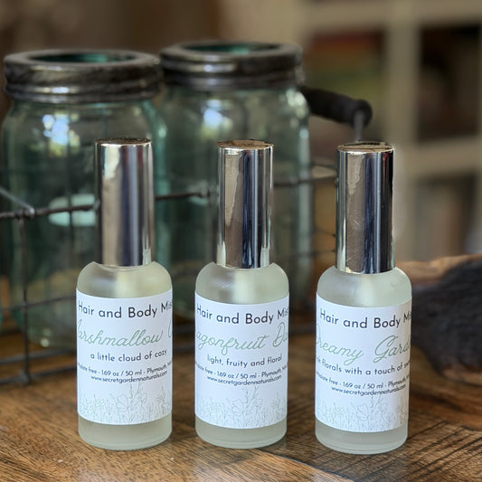 3 glass spray bottles of Secret Garden Naturals Hair and Body Mist on a wooden surface with jars behind them.