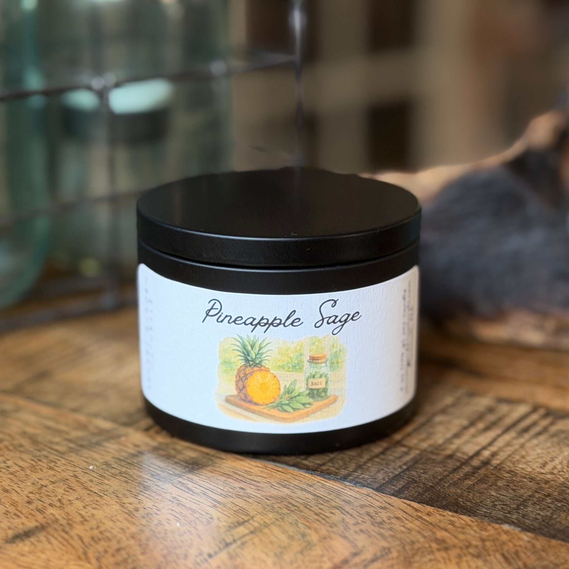 Black candle tin  labeled 'Pineapple Sage' on a wooden surface with a blurred background
