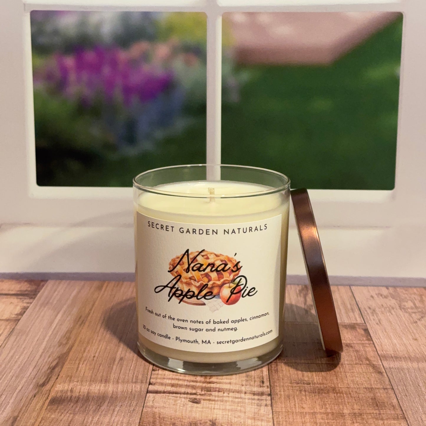 Candle labeled 'Nana's Apple Pie' on a wooden surface with a blurred window and garden view in the background.