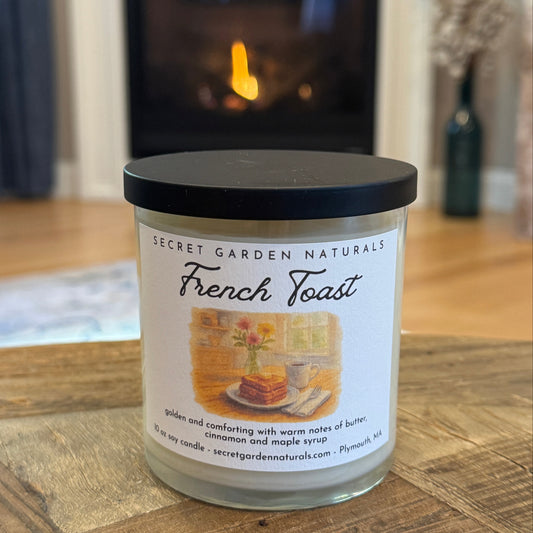 Candle labeled 'French Toast' by Secret Garden Naturals on a wooden surface with a fireplace in the background.