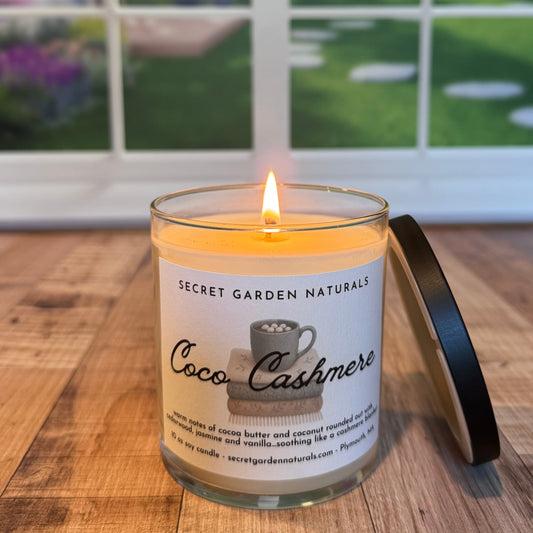 Candle labeled 'Coco Cashmere' from 'Secret Garden Naturals' on a wooden surface with a blurred outdoor background.
