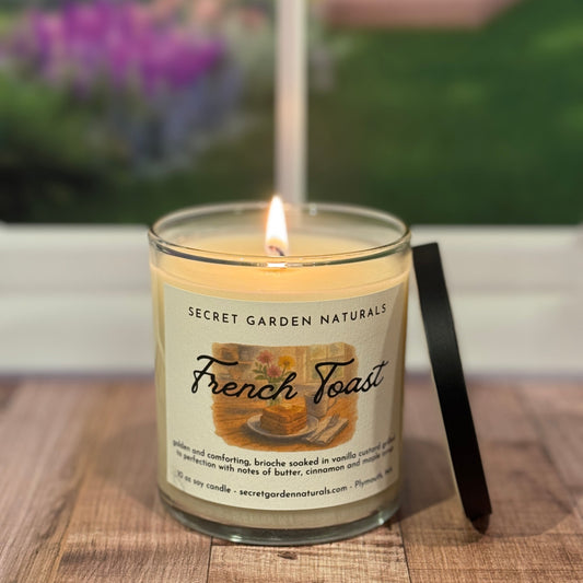 Candle labeled 'French Toast' from Secret Garden Naturals on a wooden surface with a blurred garden background.