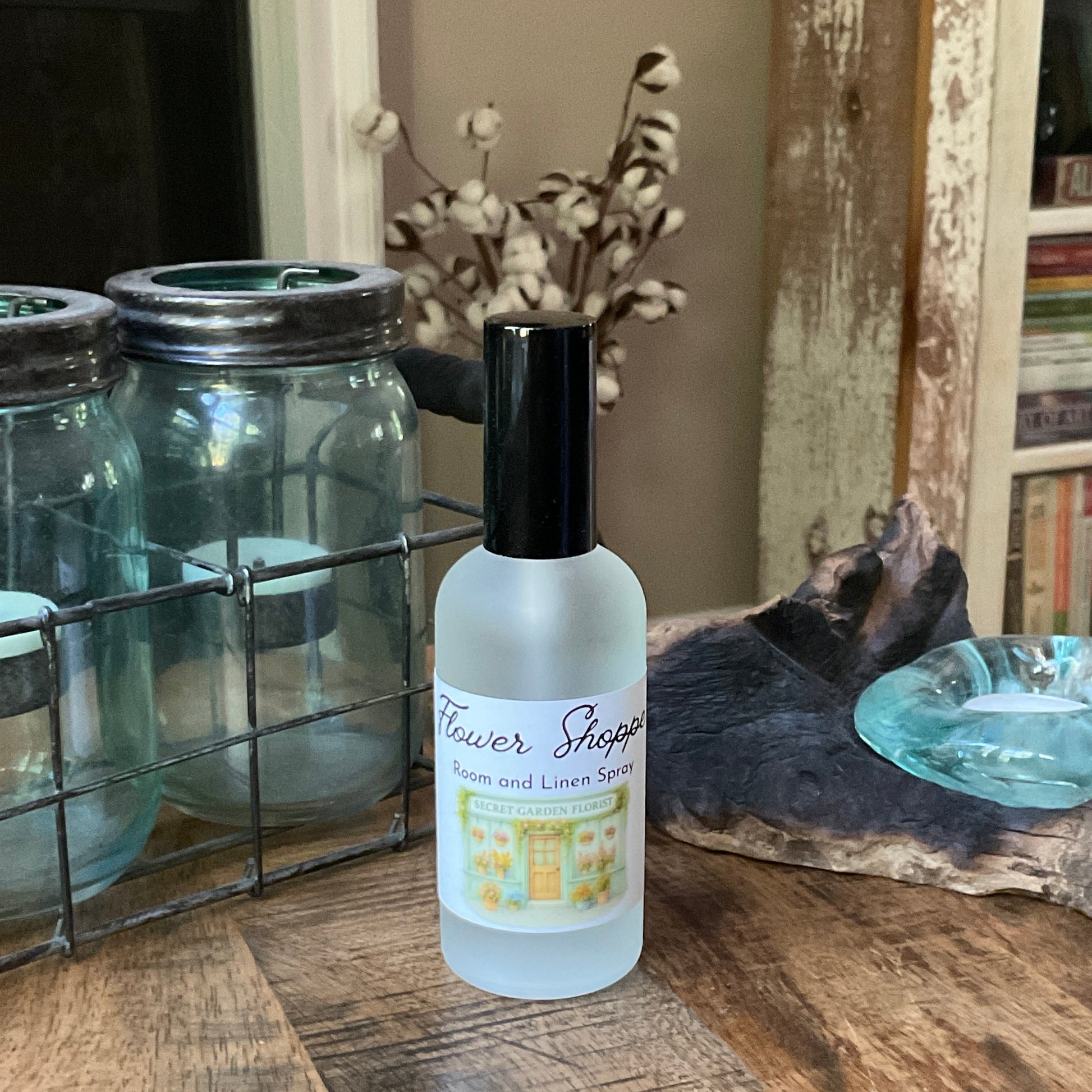 Bottle of flower shoppe room / linen spray on a wooden surface with decorative jars and books in the background.