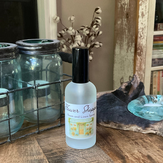 Bottle of flower shoppe room / linen spray on a wooden surface with decorative jars and books in the background.