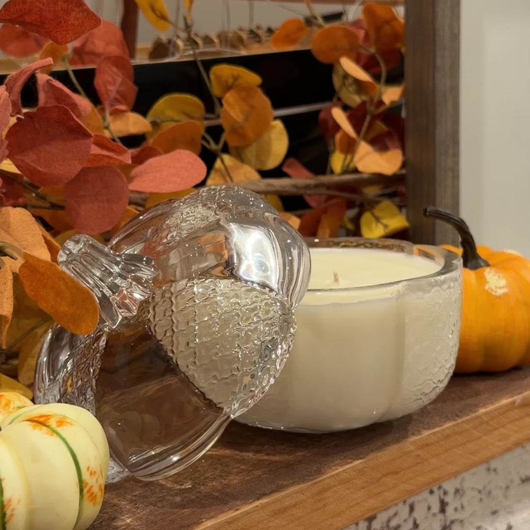 A video of a candle in a clear glass pumpkin jar on a shelf being lit surrounded by pumpkins and autumn leaves.
